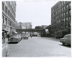 Image of cars, buildings, people crossing a street and on sidewalks, and an elevated train station. A trolley line is also visible in a cobblestone road.