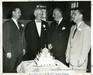 Image of four white males in suits standing above a cake inscribed with the words "15th Anniversary"