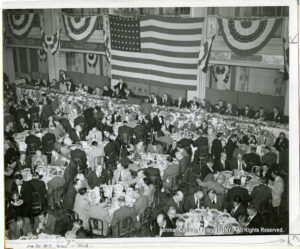 Image of American flags, and dozens of men in suits sitting at tables and eating.