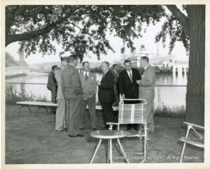 Several men in suits are standing around talking. A waterfront and a boat are visible in the background. A folding chair and table is visible in the foreground.