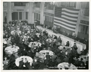 Dozens of men in suits are seated around circular tables. Image of an American flag, with a person in front of it who appears to be making a speech.