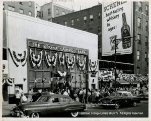Image of Bronx Savings Bank branch at 1735 University Ave. Several flags, plus cars, a 44th precinct police car, other stores, and a billboard for whiskey are visible.