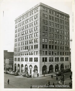 Image of a 10-story building labeled "Busher Building." The National City Bank of New York is on the first floor. Several 1940s or 1950s era cars are available, and many people cross the street. A bill board for National Cash Registers is visible above the Wool Bros. store.