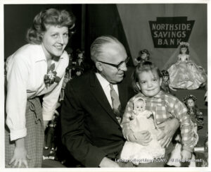 Image of a woman smiling at the camera, and a man staring at a girl holding a doll. The girl is smiling at the camera.
