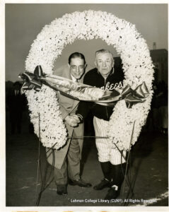 Image of Frank W. Kridel presenting Yankees manager Casey Stengel with a daffodil floral ring labeled 'Good Luck' at home plate of Yankee Stadium during opening game ceremonies.