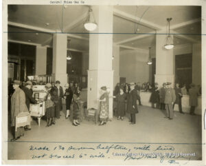 Image of a line of people. Several are looking at appliances, others appear to be paying bills. A uniformed police or security guard is also present.