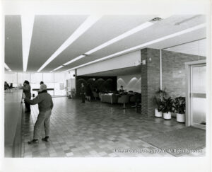 Image of several men at a bank counter. The bank has a tile floor, windows, house plants, and people seated in a waiting area.
