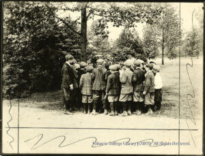 Image of a group of a dozen children clustered around a tree. Two women wearing hats, and a man in uniform stand with them. One of the women appears to be lecturing.