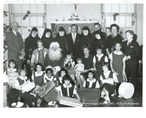 Image of about a dozen girls opening gifts. behind them are a teenage boy in a suit, a Santa Claus, several men in suits and several nuns.