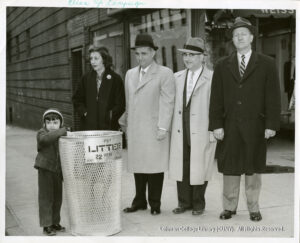Image of a child throwing something in an empty litter basket. A woman and three men stand next to him in front of the trash can, labeled "Put Litter here, District 22"