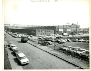 Image of a factory surrounded by parked cars, triple-parked cars and cars parked on sidewalks. A parking lot is also visible.