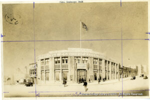 Image of an architectural drawing of a building that says "Corn Exchange Bank." Several people are walking toward the entrance. 1920s-30s era cars are visible.