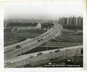 Image of a highway onramp. Several cars are visible, as is an ovular building and housing projects.
