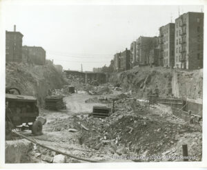 Image of construction equipment and debris in an open cut. Buildings flank the open cut.