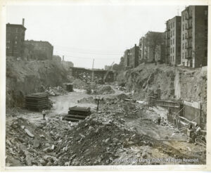 Image of debris in an open cut. Several workers walk by. Buildings flank the cut, and a bridge is visible in the background.