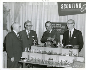 Several older men in suits looking at wire dolls, ships, and buildings