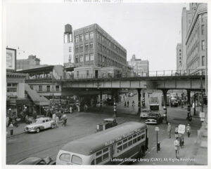 Image of a busy street scene with pedestrians, automobiles, buses, an elevated subway line, taxi and even Hearns Dept. Store employees on strike.