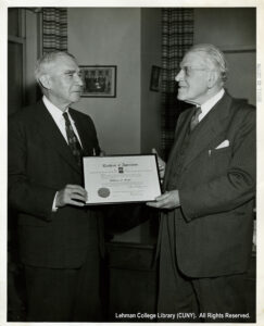 Image of one older white man in a suit handing a certificate to another older white man in a suit.