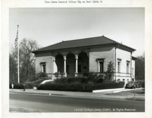 Image of a small building with two columns. An American flag, a pedestrian, and two cars are visible.