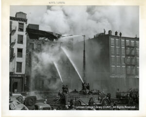Image of a building on fire. A fire truck with several fire fighters spray water at it.