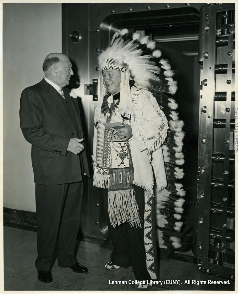 Image of a man in traditional native American attire smiles next to a bald man in a suit. They are both standing in front of a bank vault.