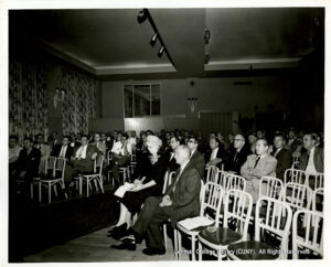 Image of mostly men in suits, as well as a woman with a note pad, sitting in chairs and staring at what appears to be a presentation.