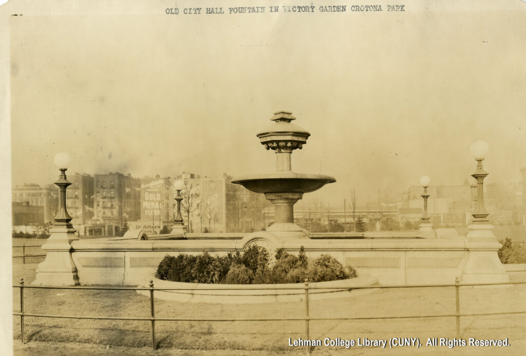 Image of a large decorative water fountain with bushes in the front, and lamp posts on the side. A nearby building is decorated with an advertisement for Gold Medal Flour.