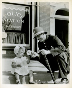 Image of a uniformed police officer with a helmet and baton handing a candy or lollipop to a small child in a white frock and bonnet.