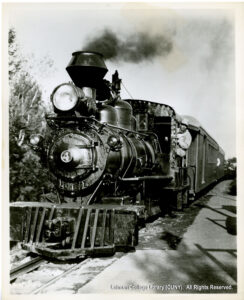 Image of a railroad engine with steam coming out. It is numbered "4" and has an engineer sticking out the side and looking at the camera.