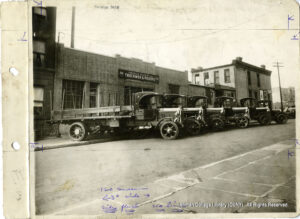 Image of several cars "combat parked" on the sidewalk outside a brick garage labeled "George Bahr Truckmen and Riggers"