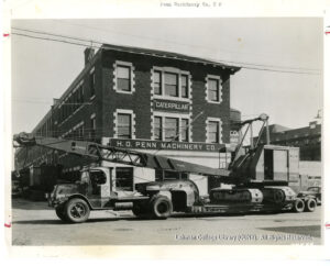 Image of a large hauling truck with a crane on its trailer in front of a brick building with sign saying "Caterpillar" and "H.O." Penn Machinery Co.