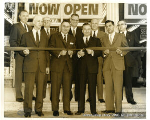 Image of several men in suits cutting a ribbon. In the background, a sign says "now open."