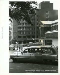 Image of cars in front of a hospital. Several people are also walking nearby, and a lamppost and tree are also visible.