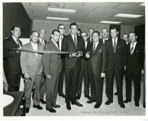 Image of many men in suits looking at a camera. The tallest one is cutting a ribbon.