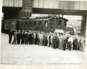 Many men in straw hats and suits in front of an electric railcar. The railcar says "New Haven 0100 Maine Bullet" and also has a box car attached.