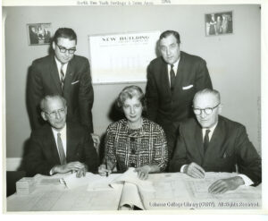 Image of several men and one woman in suits looking at architectural plans. A sign in the background says "new building."