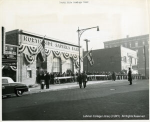 Image of a line of people with police barricades. An officer watches the crowd, and there are American flags and red-white-and-blue decorations on the bank building.