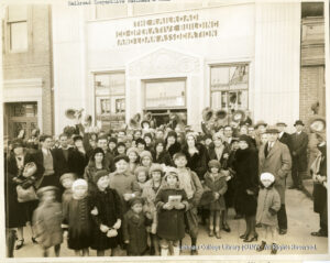 Image of many men, women, and children. Several men are tipping their hats. The building says "The Railroad Co-Operative Buidling and Loan Association"