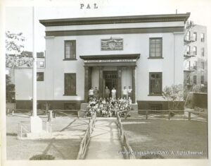 Image of several men standing and dozens of boys and girls sitting. A sign says "Police Athletic League" and "Edward P. Lynch Center".