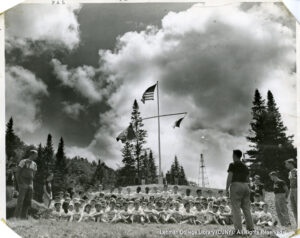 Image of several men in PAL t-shirts looking at many bosy in white t-shirts. Several flags, including an American flag, are visible, as are many evergreen trees.