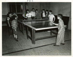 Image of two boys playing ping-pong while several others stand around watching.