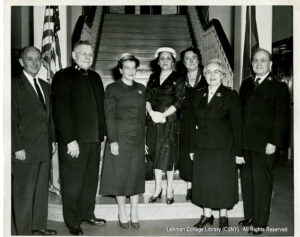 Image of several older women looking at the camera in front of a staircase. One wears a Salvation Army uniform. Two men in salvation army uniforms are also visible, as is one man in a suit. An American flag is visible in the background.