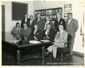 Image about 12 men in suits looking at a camera. Six are standing and six are seated at an ornate wooden table.