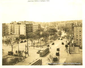 Image of apartment buildings, medians with trees, sidewalks with people, streetcars, and 1920s-era automobiles.