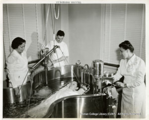 A women on a stretcher is lowered into a meal tub full of bubbling water. She is flanked by a man and two women dressed in white.