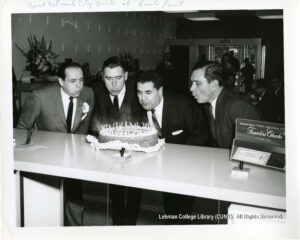 Four men blow candles out on a birthday cake next to a display of Traveler's Checks.