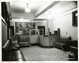 Image of a waiting rom. Leather couches, overhead lights, a reception desk, a flower, and a painting are visible.