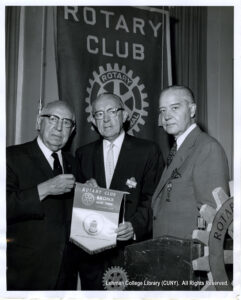Three men in suits hold a small flag that says "Rotary Club Bronx New York"