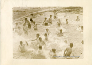 About two dozen boys wearing swim trunks splahs in water.