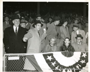 Image of many men in trench coats. one is Mayor Laguardia, who is next to two women and another two men.